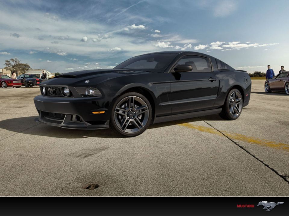 Black coupe with a hood scoop parked on pavement, other cars and people in background