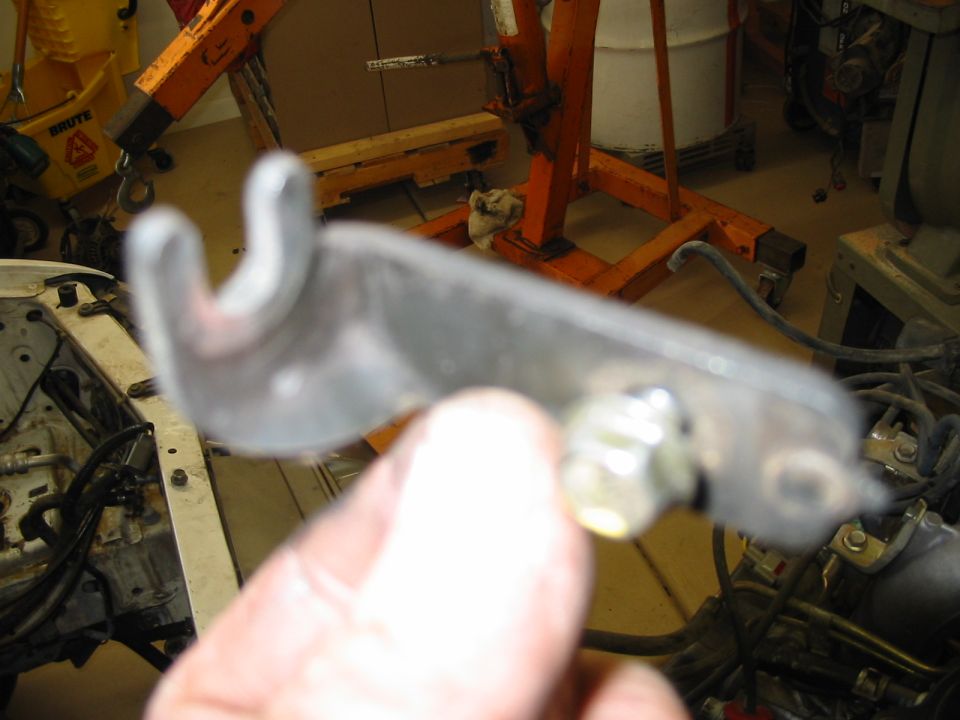 Hand holding a stamped steel bracket with a forked end in front of an orange engine hoist