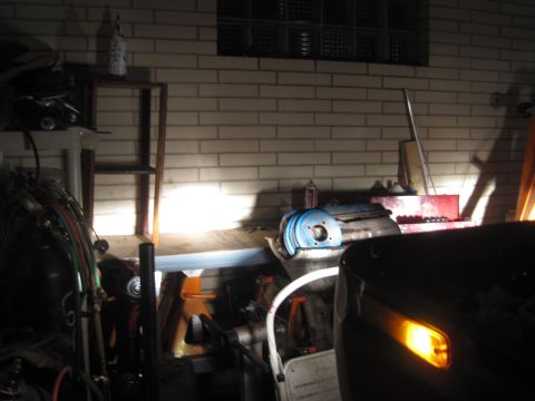 1979 Ford Bronco - A car's front corner with its headlight illuminated, set against a backdrop of garage clutter and brick wall.