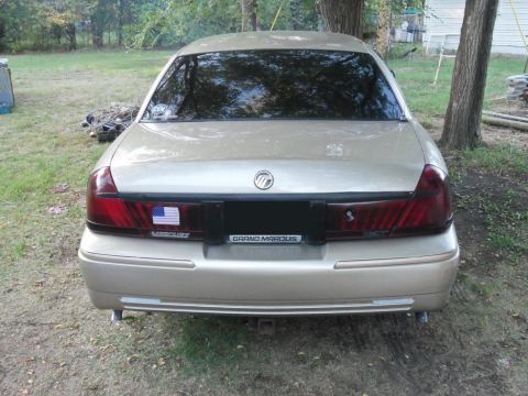 2000 Mercury Grand Marquis - The rear of a gold Mercury Grand Marquis is shown parked outdoors with trees and a house in the background.
