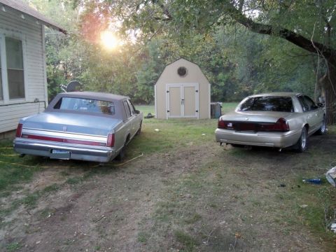 2000 Mercury Grand Marquis - Two sedans are parked in a grassy yard with a shed in the background.
