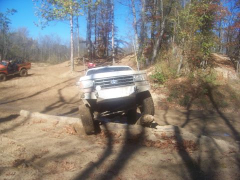 1989 Ford Bronco - A white Ford F-Series truck navigates an obstacle on a wooded trail, with another vehicle visible in the distance.