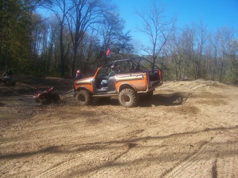 1989 Ford Bronco - Off-road vehicle with roll cage navigating a dirt track with trees and blue sky.