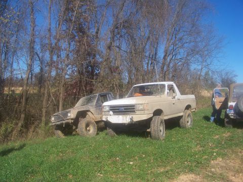 1989 Ford Bronco - Two off-road vehicles, a 1989 Ford Bronco and a Jeep Cherokee, are parked on a grassy slope with trees behind them.
