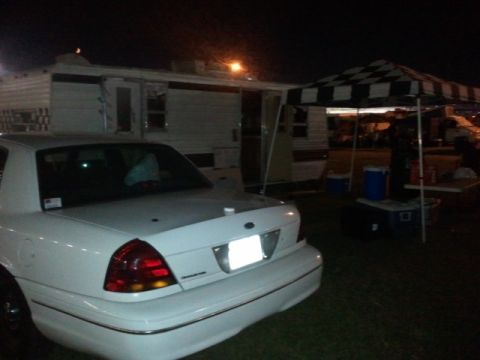 1998 Ford Crown Victoria - A white 1998 Ford Crown Victoria is parked at night near a trailer and a checkered canopy.