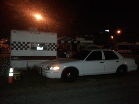 1998 Ford Crown Victoria - A white 1998 Ford Crown Victoria sedan is parked at night, illuminated by a street light and the trailer it is next to.