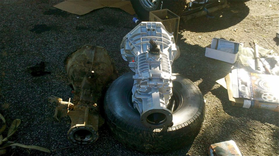 Two vehicle transmissions, one silver and one older brown, sit on the ground next to a tire. Boxes and other parts are visible in the background