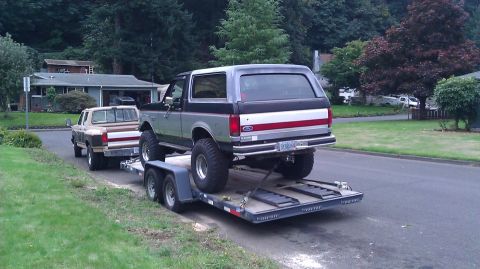 1989 Ford Bronco - A silver and black Ford Bronco is loaded onto a trailer, with a tan pickup truck visible behind it.