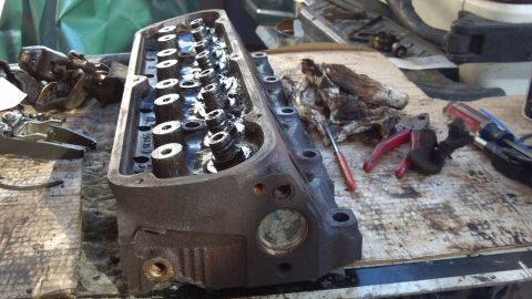 1990 Ford Bronco - A disassembled engine cylinder head sits on a cluttered workbench, surrounded by tools and oily rags.