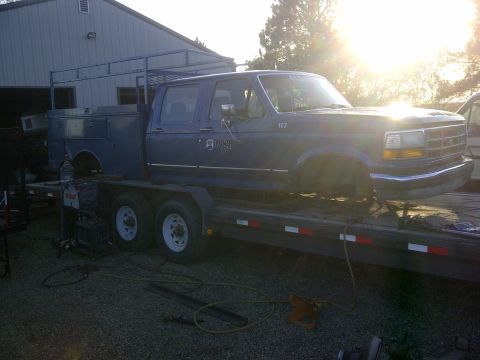 1979 Ford E250 - A blue Ford F-Series truck with a utility bed and rack is transported on a trailer, missing its front wheels.