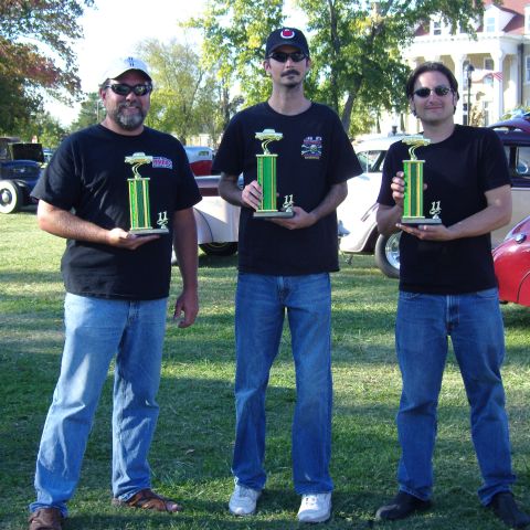 2007 Ford Mustang - Three men proudly display their trophies at an outdoor event with vintage cars visible.