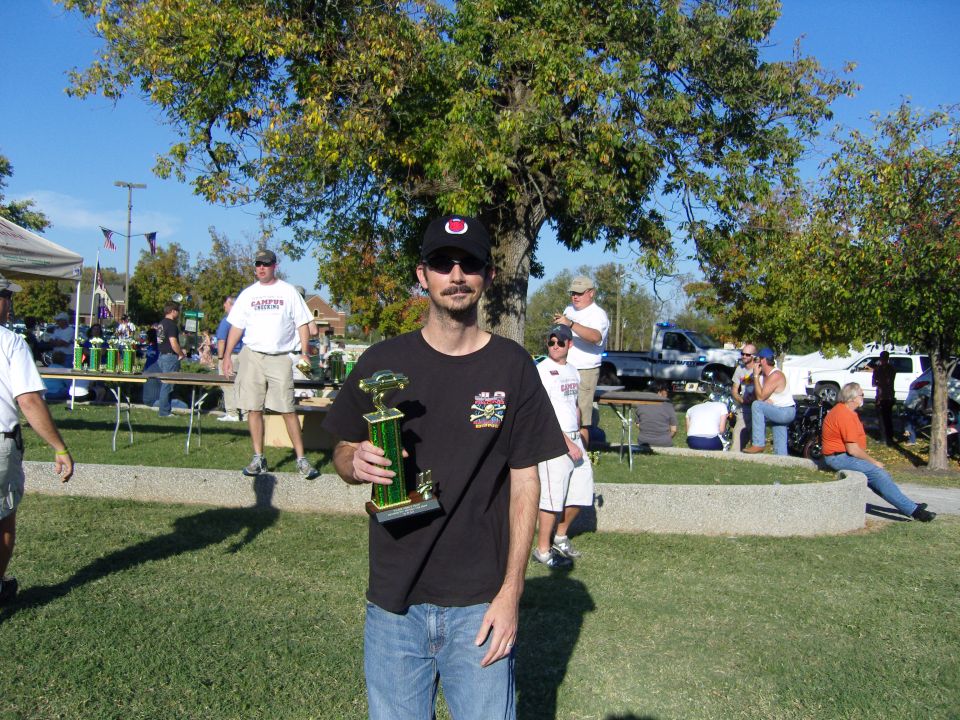 guy with trophy, other people around, tables with trophies, cars in background