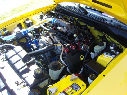 2007 Ford Mustang - The engine bay of a yellow 2007 Ford Mustang, aftermarket blue intake piping and a yellow Optima battery.