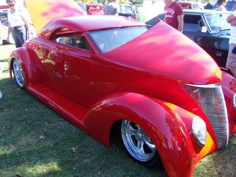 2007 Ford Mustang - A bright red custom coupe hot rod with its hood open, displaying a chrome grille and custom wheels at an outdoor car show.