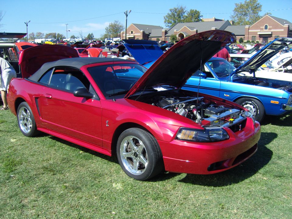 Red 2007 Ford Mustang convertible with hood open, parked on grass at car show
