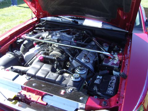 2007 Ford Mustang - The engine bay of a red car is open, revealing the V8 engine and its components under a sunny sky.