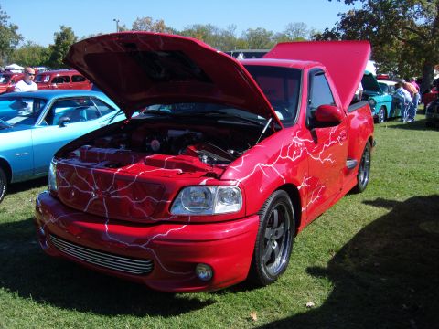 2007 Ford Mustang - A red pickup truck with custom lightning bolt graphics is displayed with its hood open at an outdoor car show.