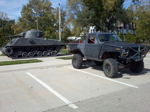 1986 Ford Bronco - A heavily modified pickup truck is parked next to a military tank in a parking lot.
