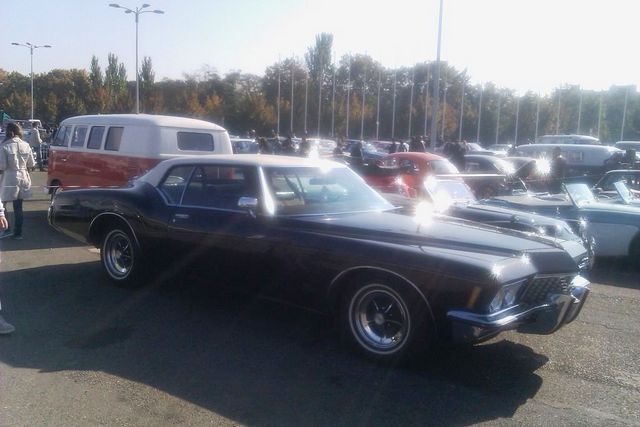 Dark coupe with white roof, likely a 1973 Buick Riviera, parked outdoors. Red and white van visible to the left
