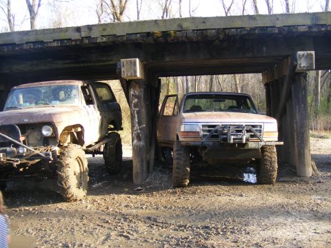 1978 Ford Bronco - Two modified trucks are parked under a wooden overpass in a wooded area.