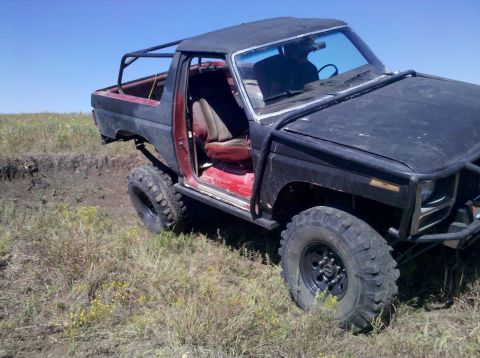 1986 Ford Bronco - This heavily modified SUV, possibly a Ford Bronco, sits off-road with its door open and a prominent roll cage.