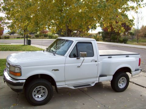 1993 Ford F-150 - This white Ford F-150 pickup truck is parked on a paved surface with trees and a residential area visible.