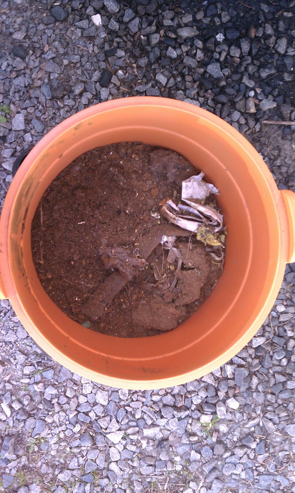 Orange bucket filled with dirt, leaves, and trash on a gravel surface