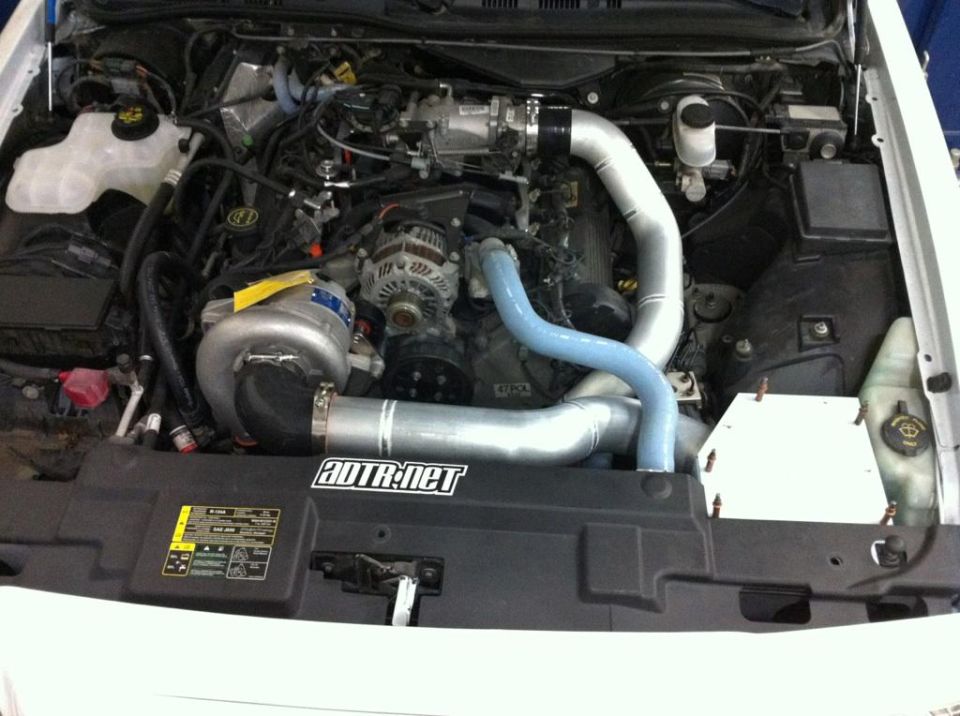 Engine bay with a large silver supercharger and custom piping. ADTR.net sticker visible