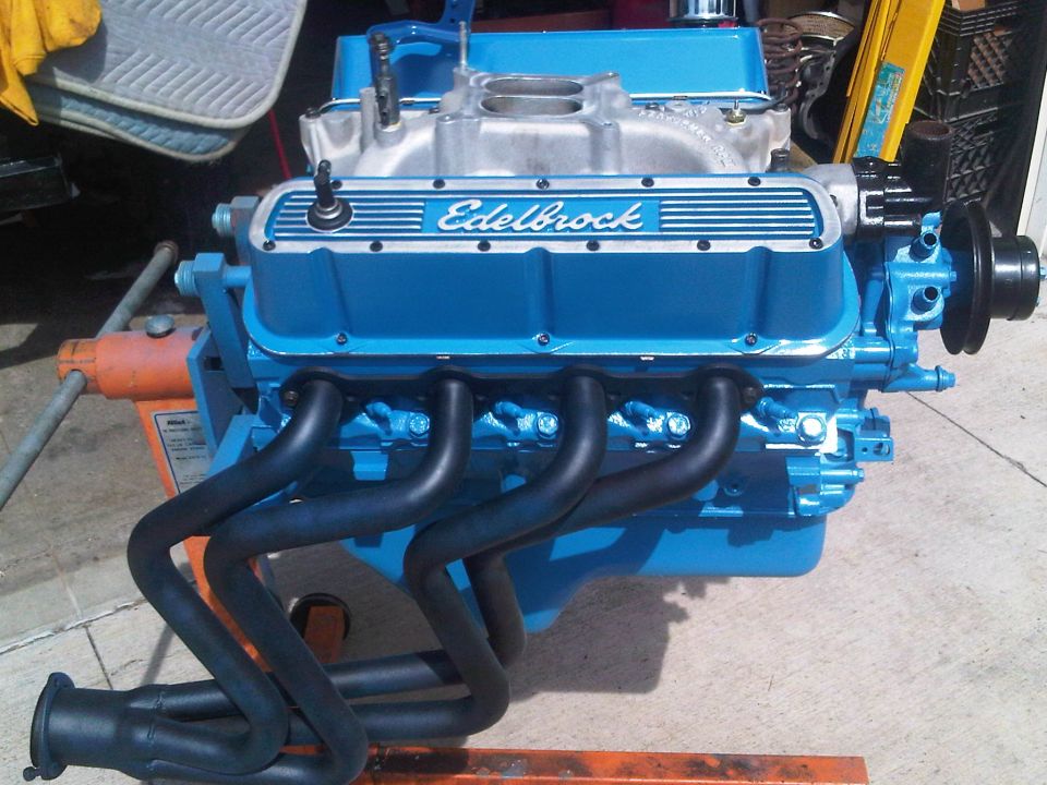 Blue Edelbrock V8 engine with black headers and intake manifold. Engine is on an orange stand