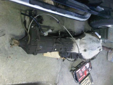 1985 Ford Bronco - A vehicle transmission assembly is shown on a concrete surface, with parts of an exhaust pipe and a tire in the background.