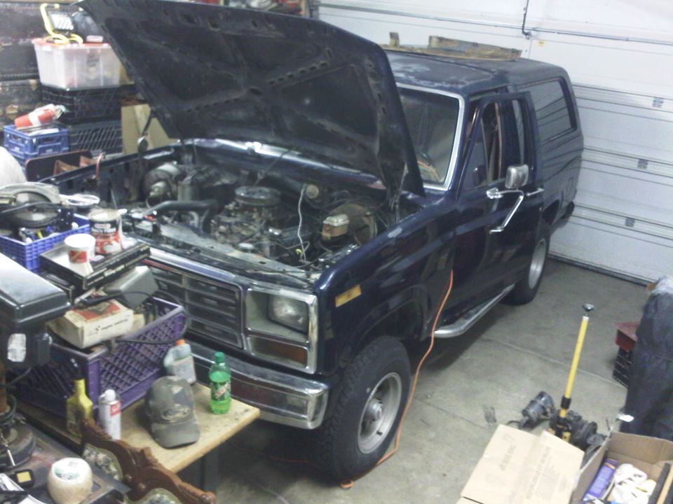 Dark blue Ford Bronco with hood open, showing engine. Parked in a garage with tools and storage around