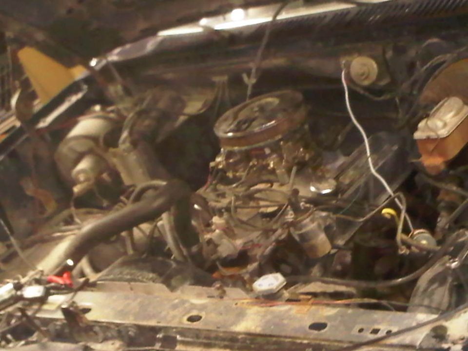 Engine bay with a V8 engine, carburetor, and various hoses. The condition appears worn