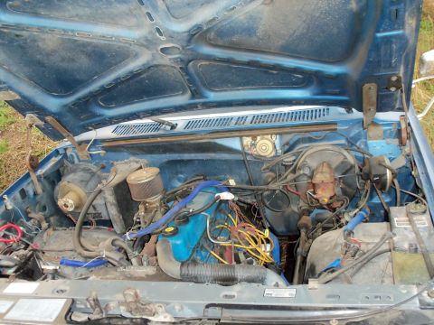 1985 Ford Bronco - The engine bay of a blue Ford truck is shown with the hood open, revealing the engine components.