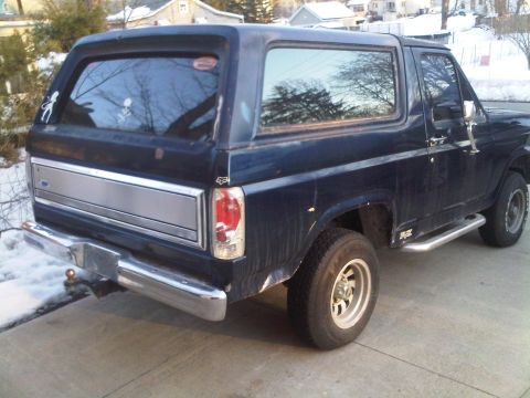 1985 Ford Bronco - The rear of a dark blue Ford Bronco is shown parked on a driveway in the snow.
