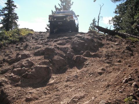 1991 Ford Bronco - A yellow off-road vehicle navigates a challenging, rocky incline under a clear blue sky.
