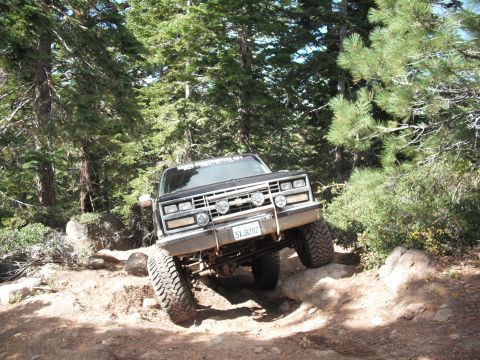 1991 Ford Bronco - This lifted pickup truck navigates a rocky off-road trail surrounded by pine trees.