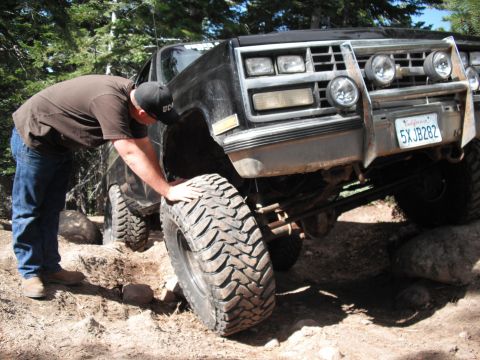 1991 Ford Bronco - Man inspects tire on a lifted truck navigating rocky terrain.