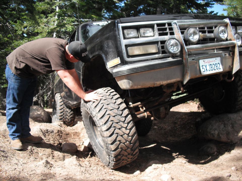 Man checks large off-road tire on lifted black truck with aftermarket bumper and lights. California plate 5XJB282