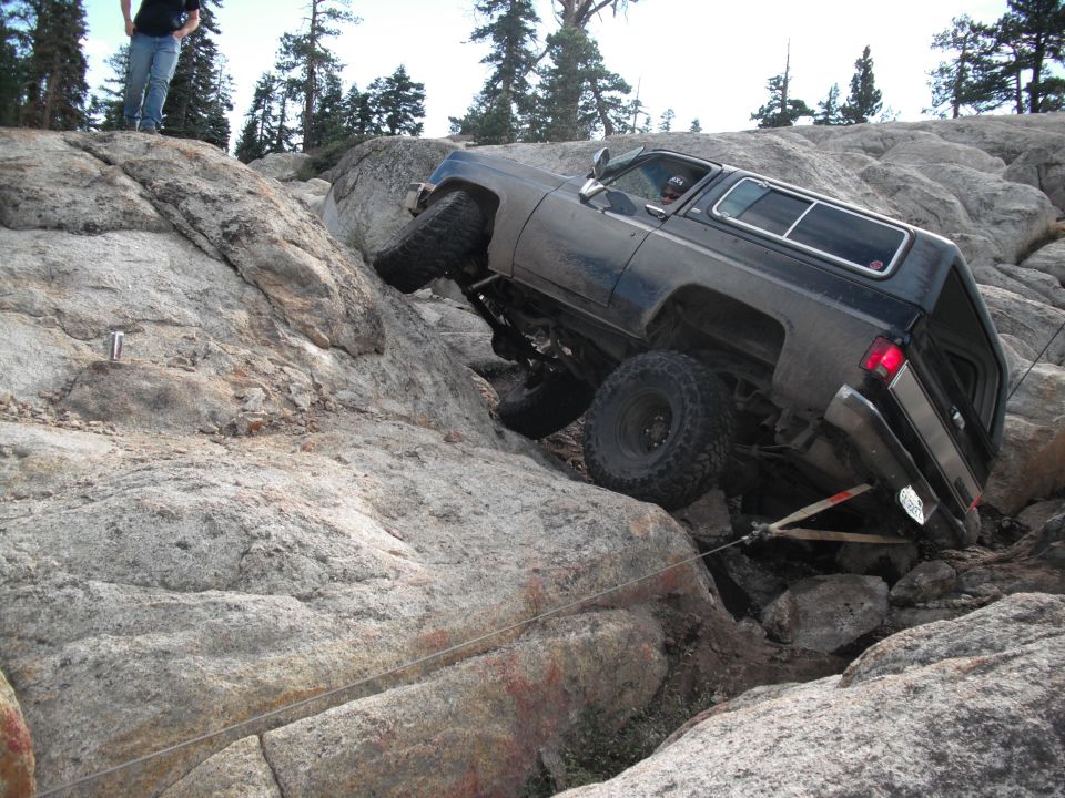 dark off-road SUV with large tires climbing over large rocks, one wheel lifted high