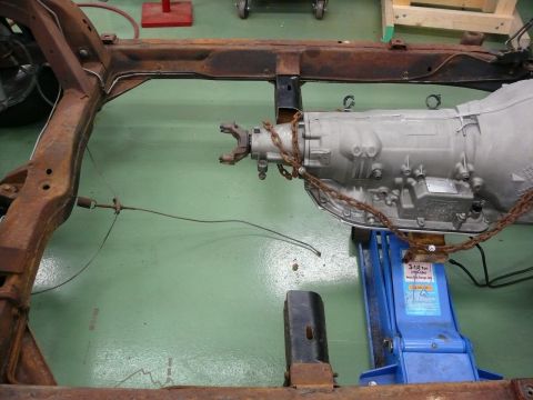 1976 Chevrolet Corvette - The rusty frame of a vehicle is shown with a transmission supported by a blue hydraulic jack.
