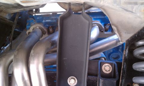 1979 Ford Bronco - Close-up view of engine components, including exhaust pipes and a blue engine block with a Ford logo on a black cover.