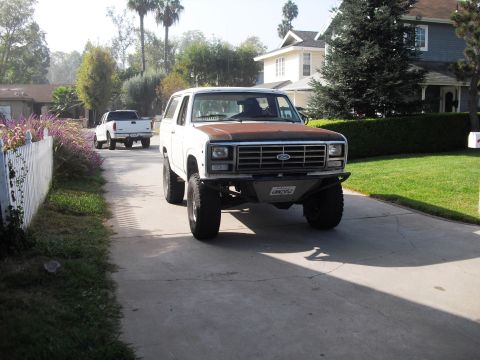 1986 Ford Bronco - A white Ford Bronco with a rusty hood drives down a suburban driveway, followed by a white pickup truck.