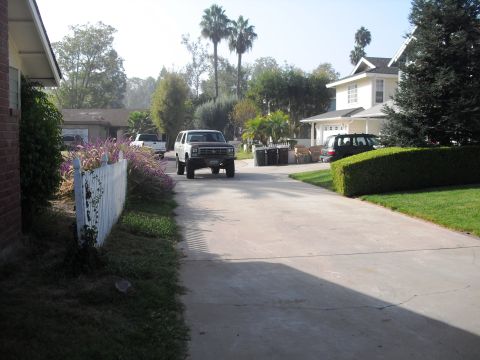 1986 Ford Bronco - A white SUV drives down a suburban driveway, passing a white picket fence and manicured hedges.