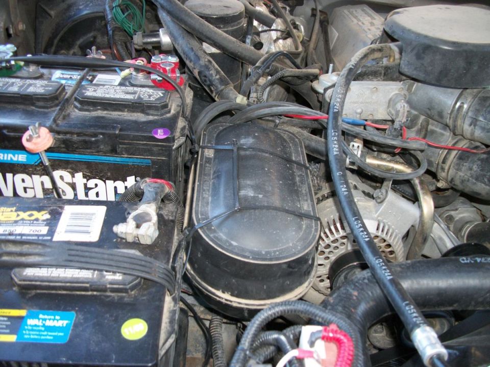 Engine bay showing a large battery, air filter housing, alternator, and various hoses and wires