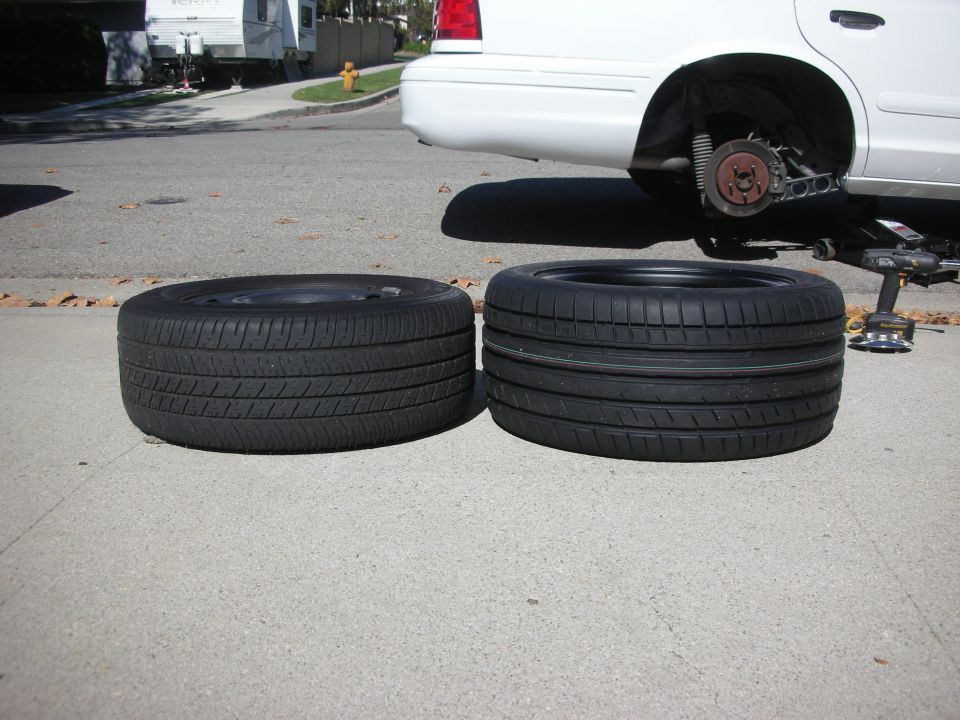 Two tires, one on a rim, next to a white 2004 Ford Crown Victoria with rear wheel removed