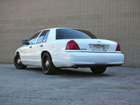 2004 Ford Crown Victoria - Rear view of a white 2004 Ford Crown Victoria sedan with black wheels parked against a textured wall.