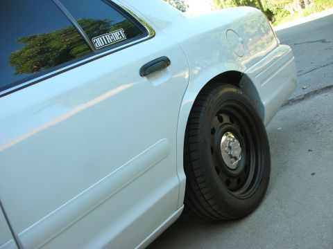 2004 Ford Crown Victoria - Rear side view of a white 2004 Ford Crown Victoria, highlighting its steel wheel and a sticker on the window.