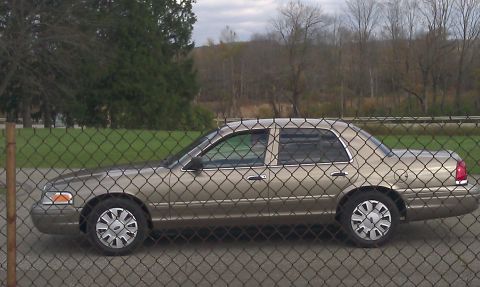 2007 Ford Crown Victoria - A tan 2007 Ford Crown Victoria is seen from the side, behind a fence with trees in the background.