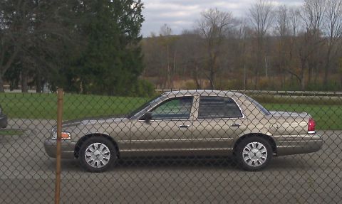 2007 Ford Crown Victoria - A gold 2007 Ford Crown Victoria is seen from the side, parked behind a fence with trees in the background.