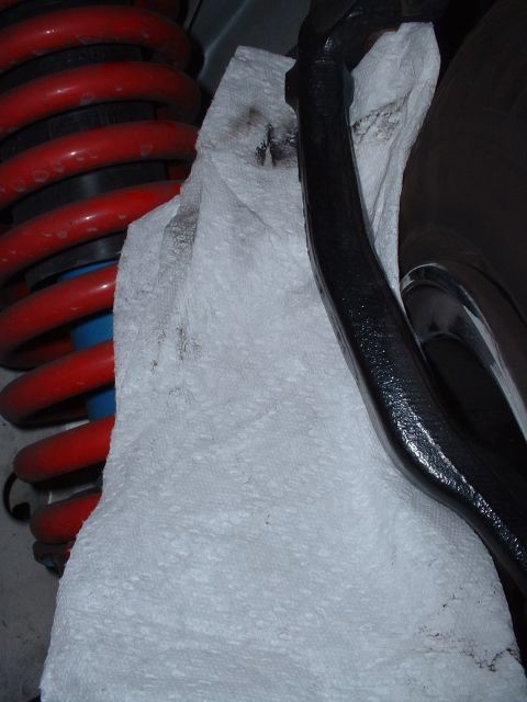 1997 Mercury Cougar - A close-up view of automotive suspension components, including a red coilover shock and a black linkage, with a paper towel absorbing fluid.